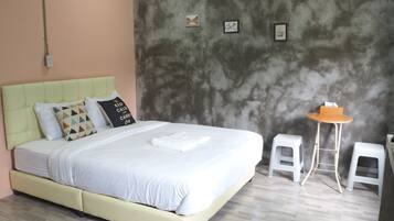 A bed with a light green headboard, white bedding, and multiple pillows. There is a small wooden table with a stool beside the bed. The room has a concrete wall with two framed pictures and a power outlet.