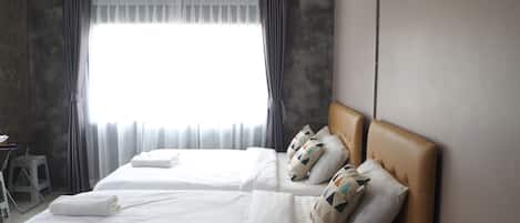 A hotel room with two beds, a window with curtains, a small table with a chair, and a wall-mounted radiator.