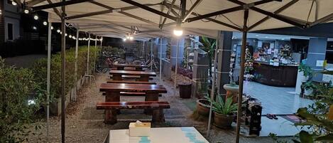 Een terras met houten tafels en banken, omgeven door plantenpotten en een overdekte constructie met lichtslingers.