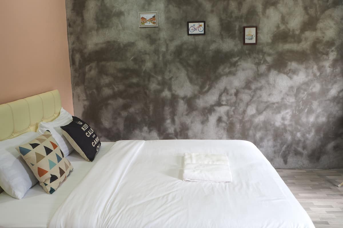 Una cama bien tendida con ropa de cama blanca y una toalla doblada, un cabecero y una pared gris texturizada con tres cuadros enmarcados.