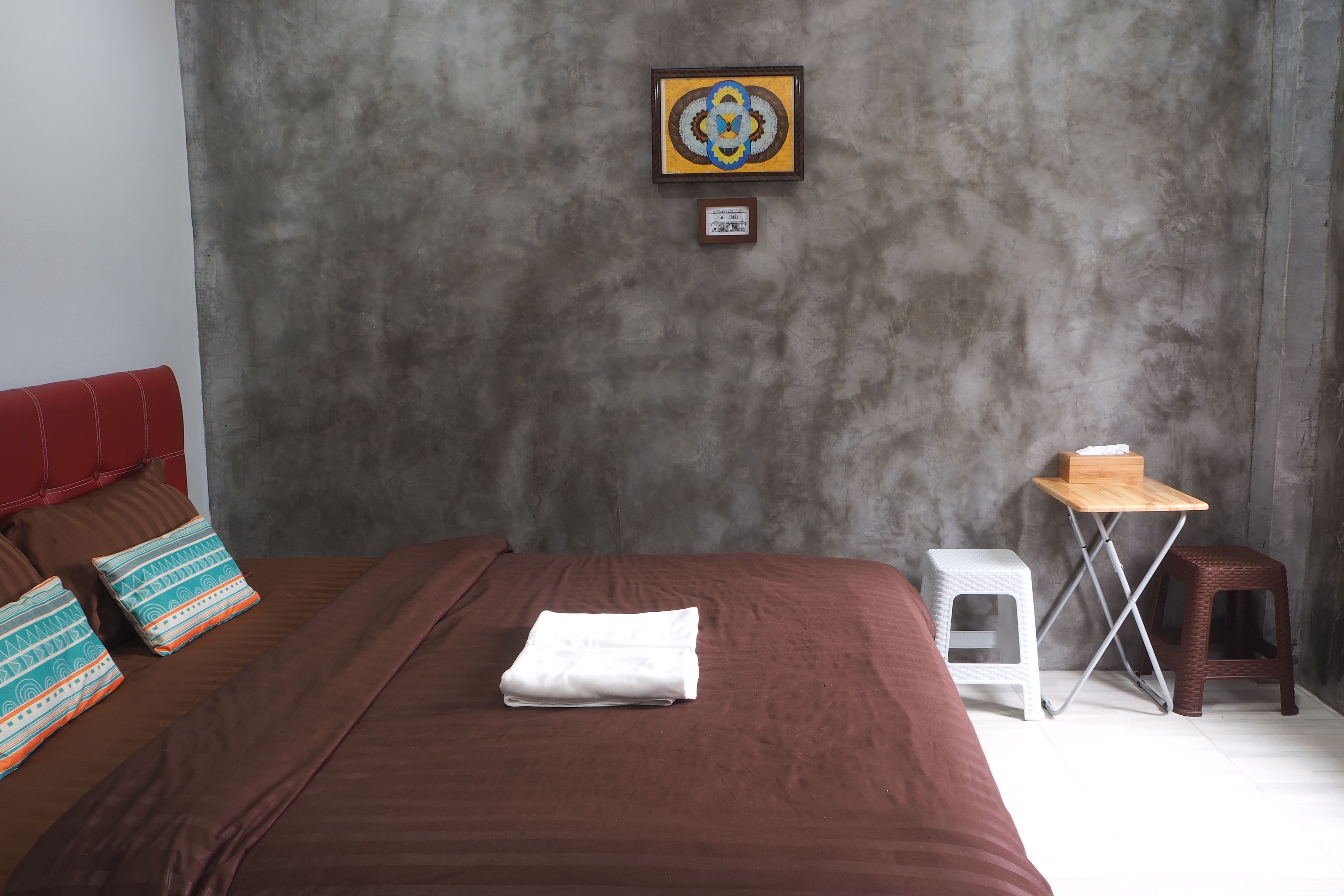 A bedroom with a large bed, a small table, two stools, and a framed picture on the wall.