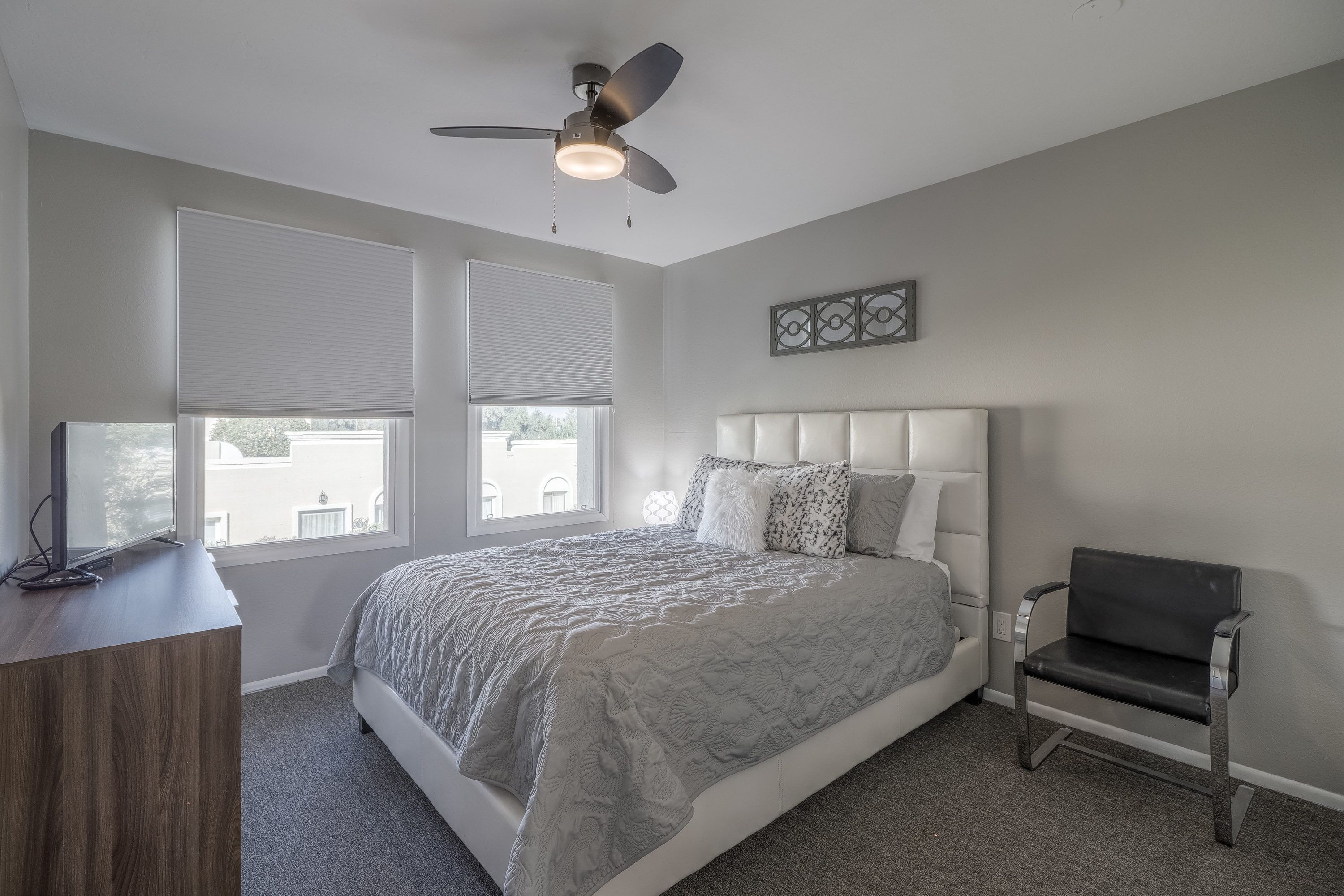 Une chambre à coucher comprenant un lit, un bureau, une chaise, un ventilateur de plafond et deux fenêtres équipées de stores.