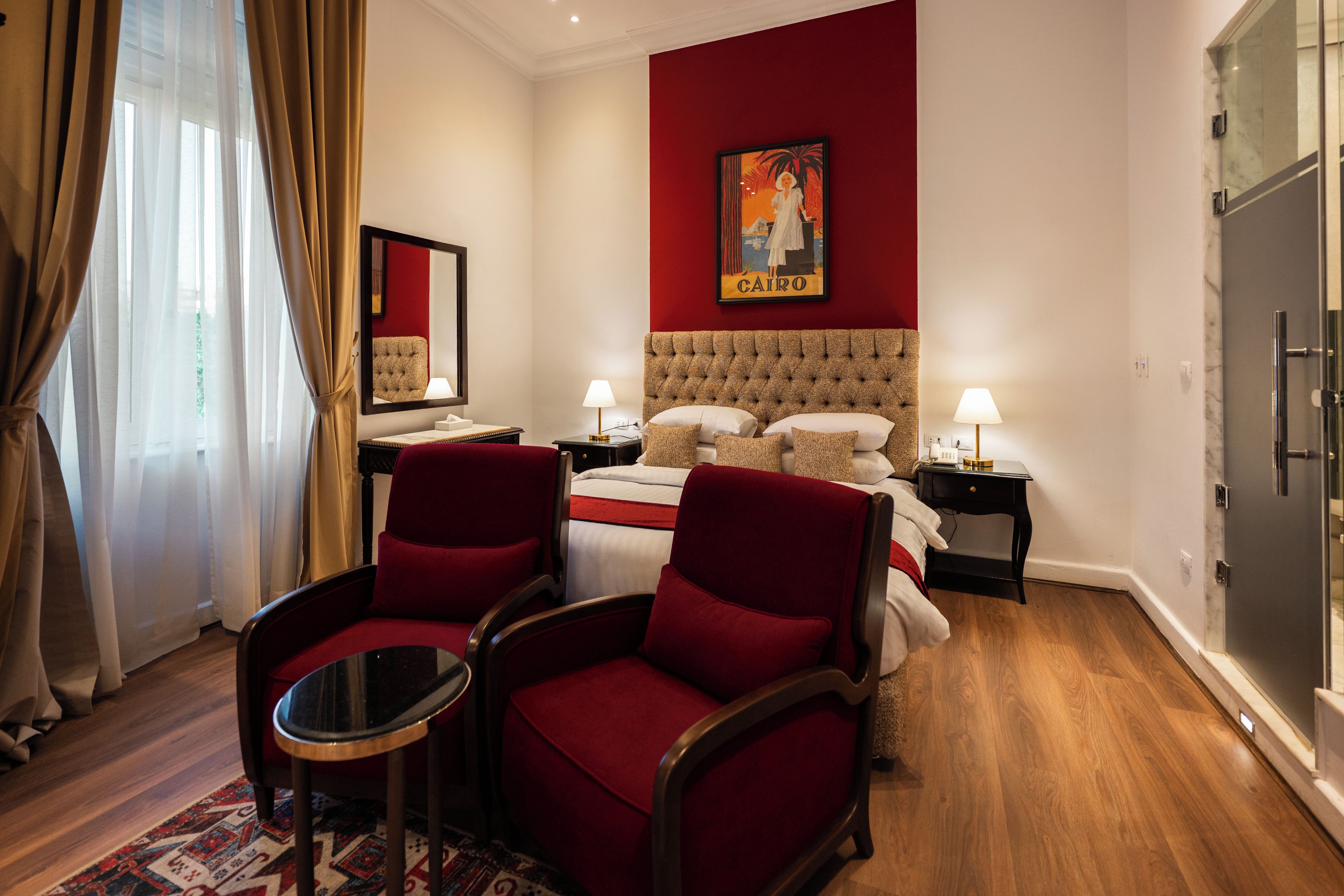 Una habitación de hotel con una cama grande, dos sillones rojos, una mesita, una mesita de noche, un cuadro y un armario.