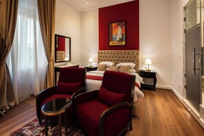 A hotel room with a large bed, two red armchairs, a small table, a nightstand, a painting, and a wardrobe. - 1920s Boutique Hotel and Restaurants (Cairo)