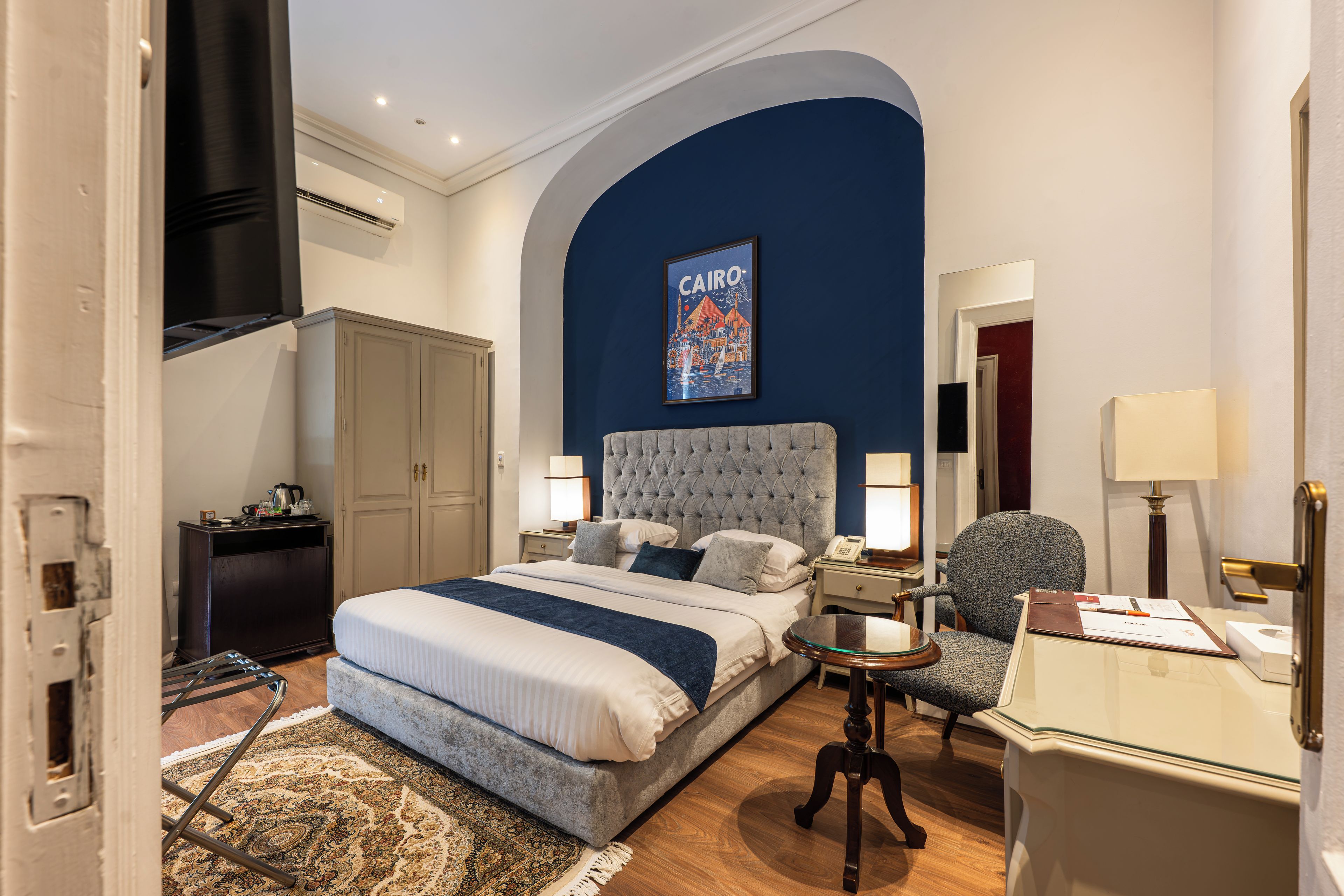 Una habitación de hotel con cama, escritorio, silla, armario y un cuadro de El Cairo.
