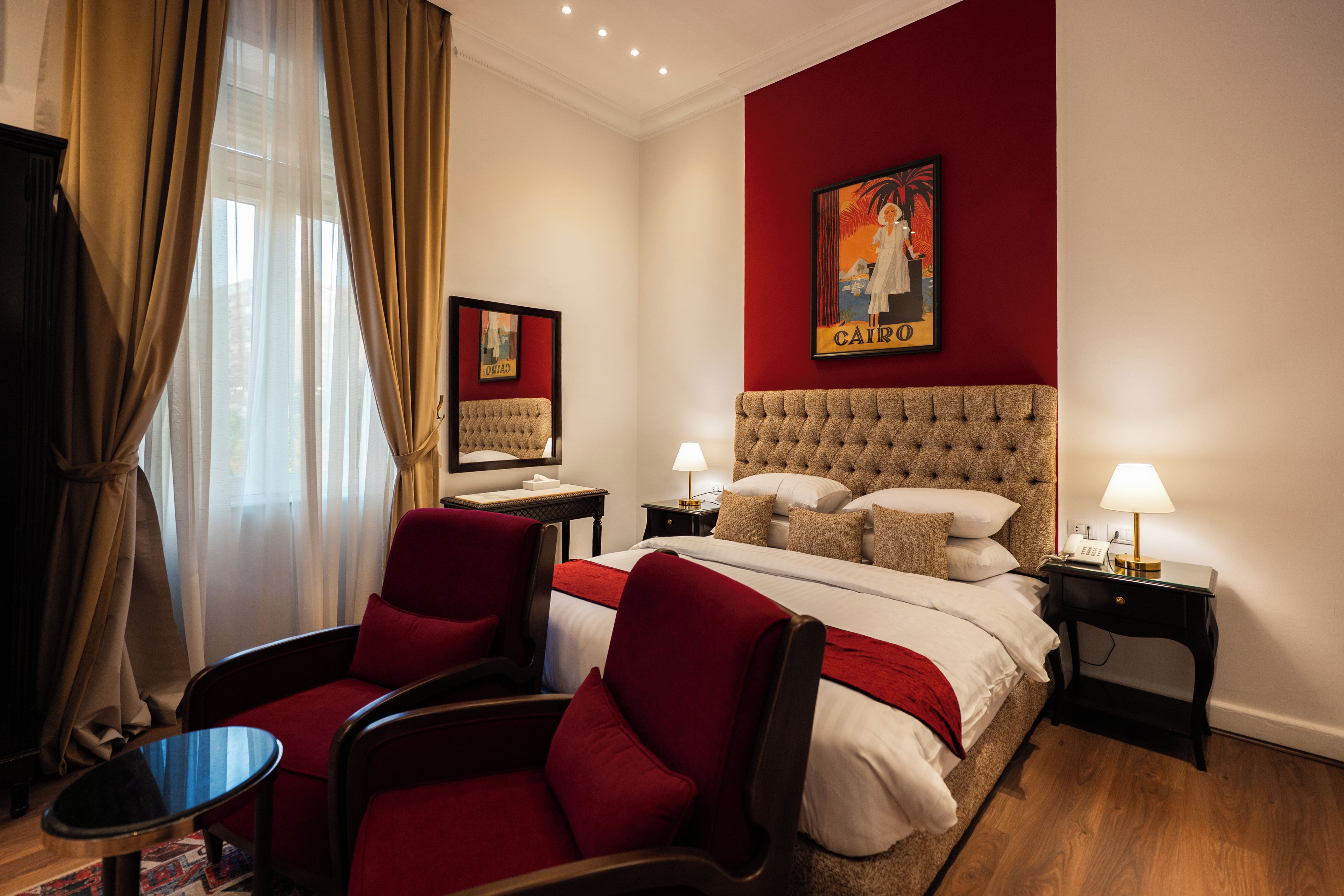 Ein Hotelzimmer mit einem Bett, zwei Nachttischen, einem Schreibtisch, zwei Stühlen, einer roten Wand und einem Fenster mit Vorhängen.