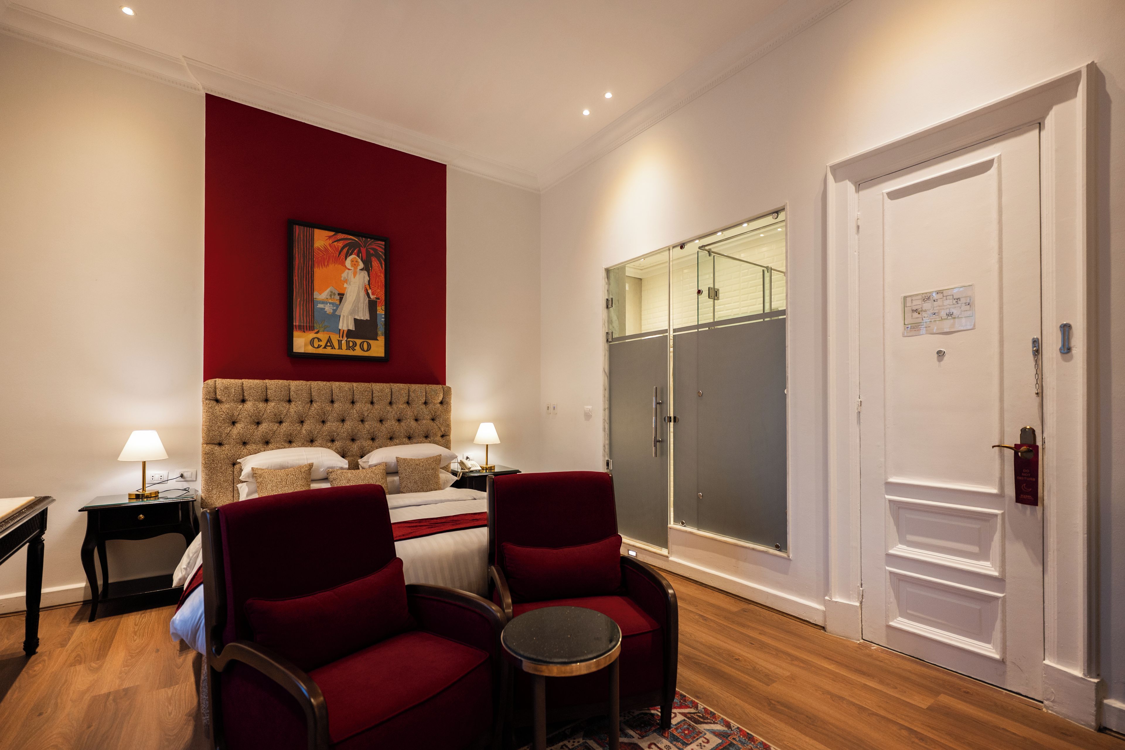 Une chambre d’hôtel comprenant un lit, deux tables de chevet, un fauteuil rouge, une petite table ronde, une armoire et une porte dotée d’une serrure à carte magnétique.