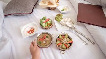 Un desayuno ordenado sobre una cama con un almohadón, un tazón de sopa, una ensalada, un plato de aceitunas y una bandeja de pan.