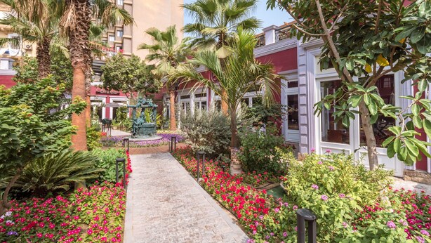 A courtyard with a central pathway, surrounded by palm trees, flowering plants, and a statue. - 1920s Boutique Hotel and Restaurants (Cairo)