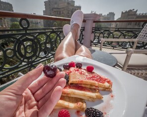 A person holding a plate with pancakes and berries, sitting on a balcony with a railing and buildings in the background. - 1920s Boutique Hotel and Restaurants (Cairo)
