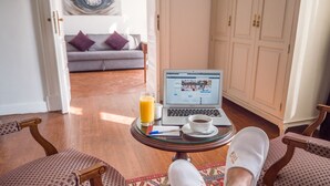A room with a laptop on a small table, a glass of orange juice, and a cup of coffee. There are patterned chairs and a sofa with cushions. A painting is on the wall, and a wardrobe is visible. - 1920s Boutique Hotel and Restaurants (Cairo)