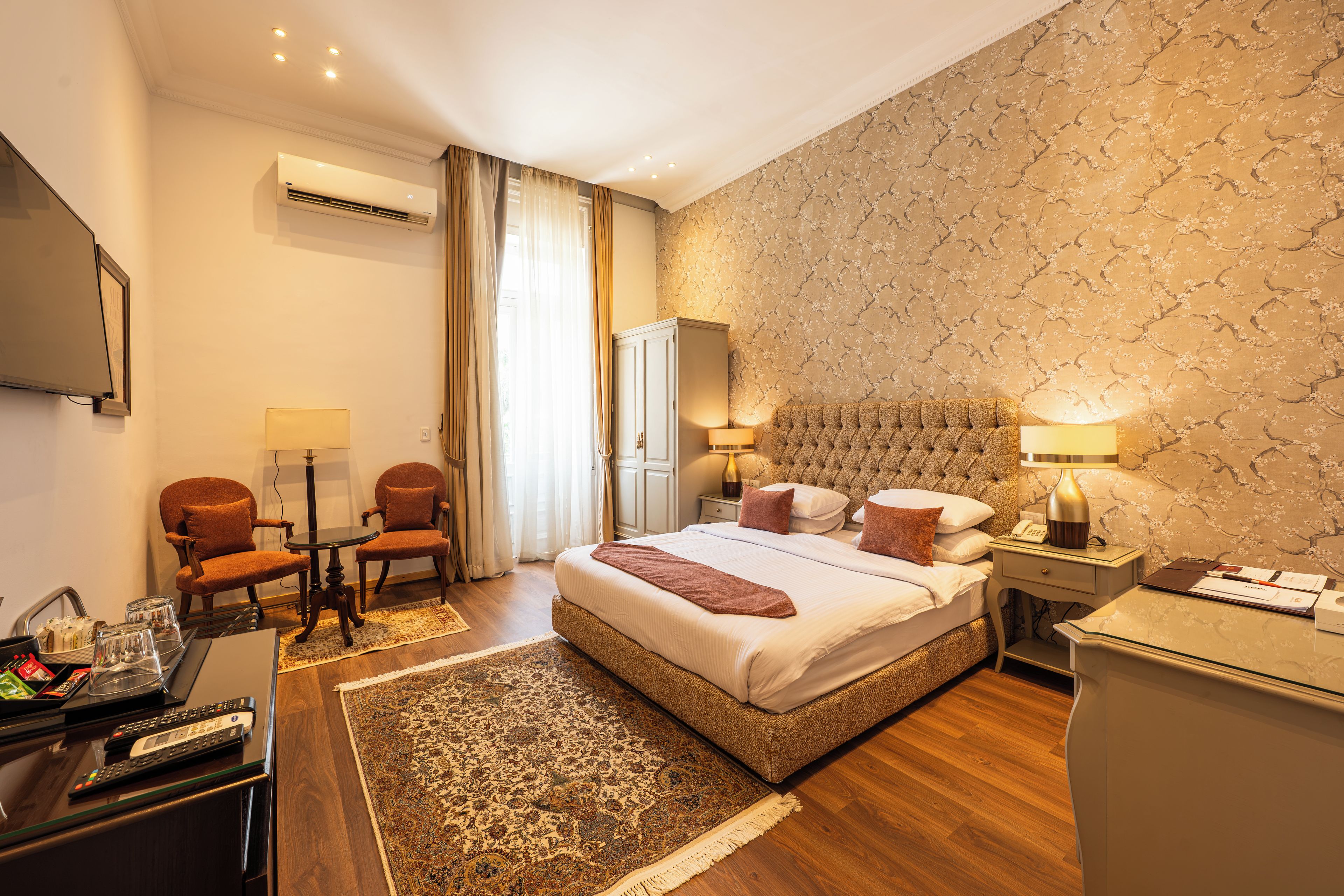 A hotel room with a bed, two armchairs, a desk with a lamp, a nightstand, and a wall-mounted TV.