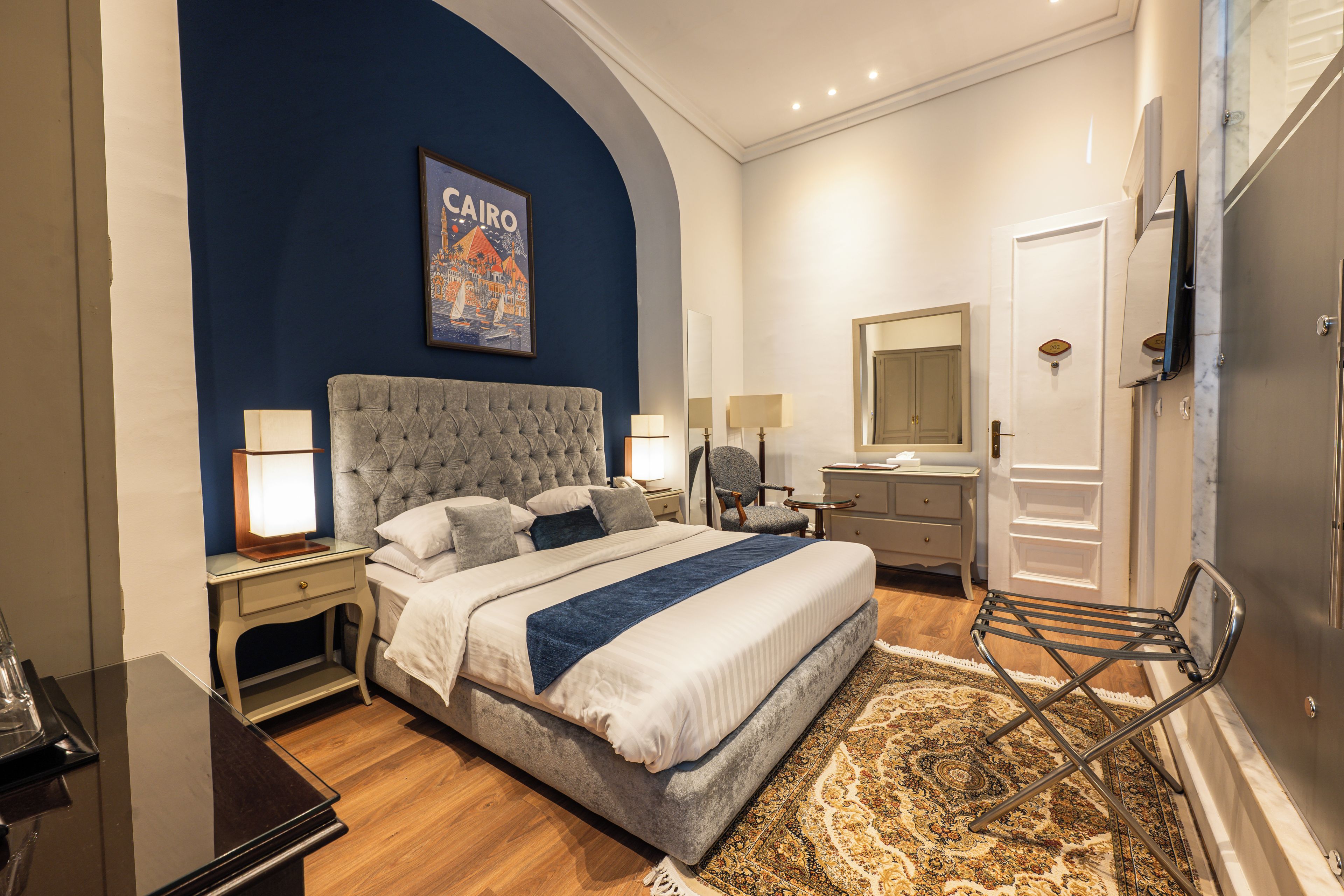 Habitación de hotel con cama, l ámparas de noche, mesita de noche, silla, cómoda, espejo y puerta con número de habitación.