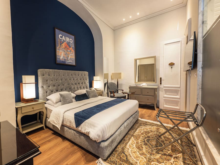 Quarto de hotel com cama, abajures, mesa de cabeceira, cadeira, cômoda, espelho e porta com número do quarto.