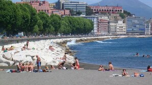 Une plage où des gens se prélassent au soleil et se baignent, un rivage rocheux et des bâtiments en arrière-plan.