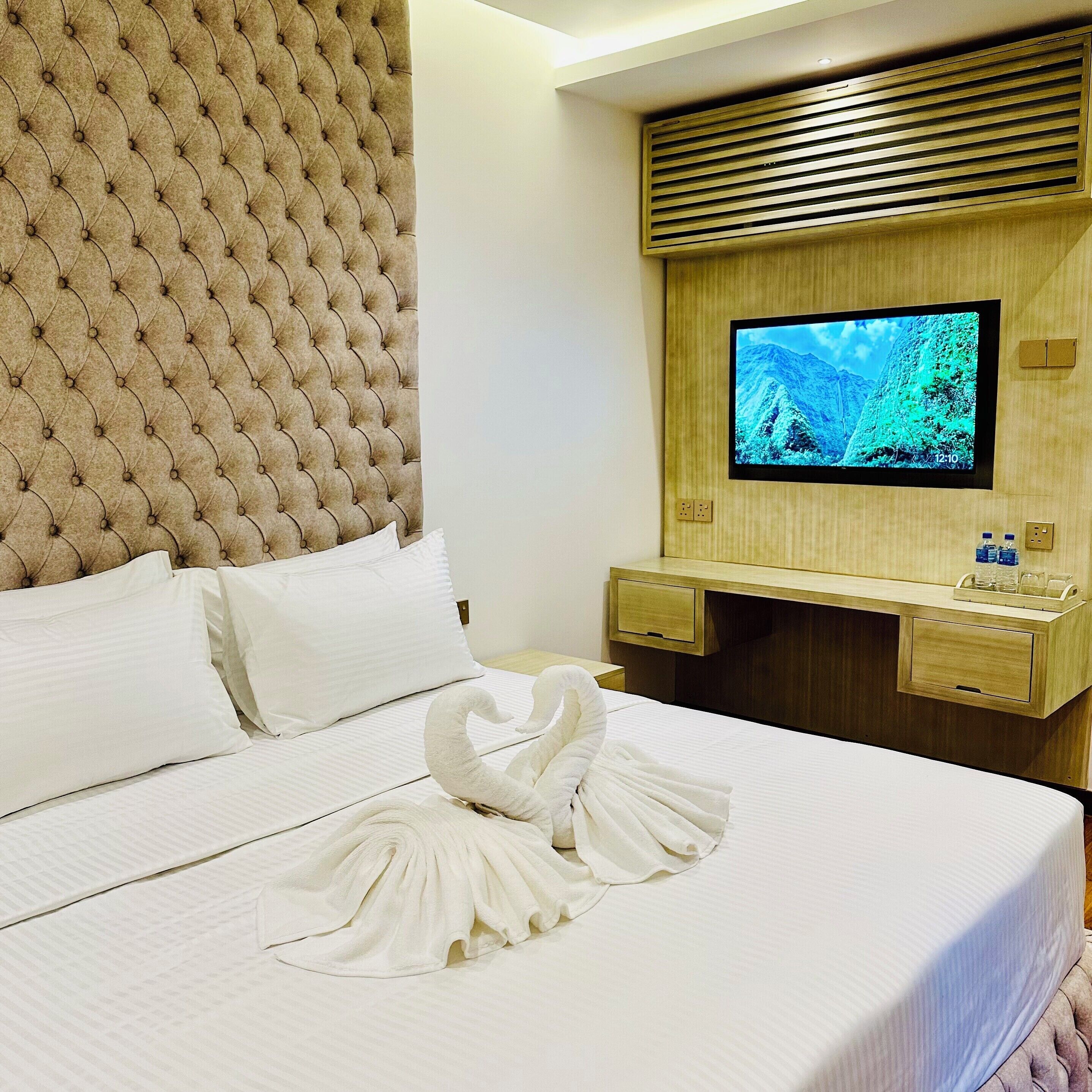 A hotel room with a tufted headboard, a flat-screen TV mounted on a wooden panel, a bed with white linens, and a swan-shaped towel on the bed.