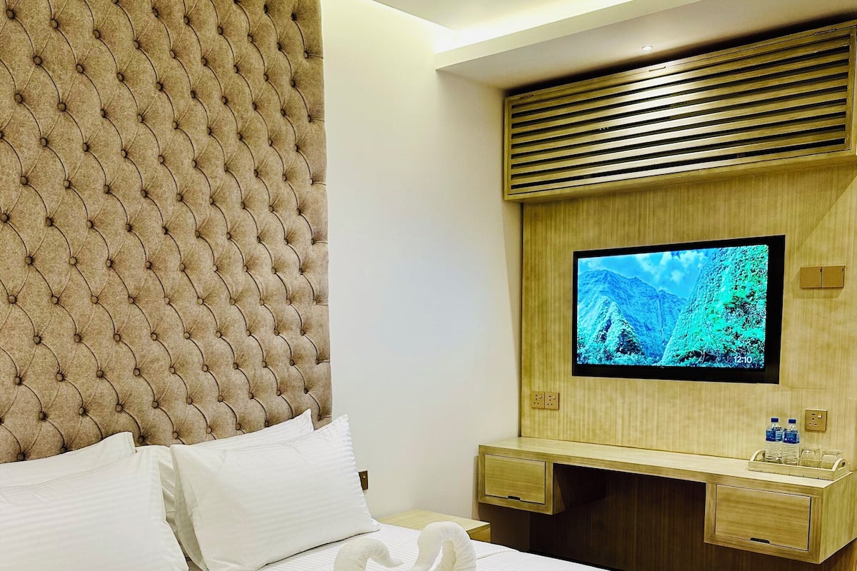 A hotel room with a tufted headboard, a flat-screen TV mounted on a wooden panel, a bed with white linens, and a swan-shaped towel on the bed.
