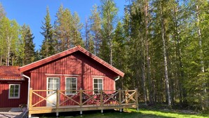 A red cabin with a wooden deck and a covered porch, surrounded by a forest.