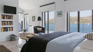 Una camera da letto moderna con un letto grande, una scrivania e una sedia. Si nota una libreria piena di libri, una TV a parete e una finestra con vista sul mare.