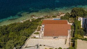 Vista aerea di una proprietà costiera con un edificio a tetto rosso, acque cristalline e vegetazione lussureggiante.