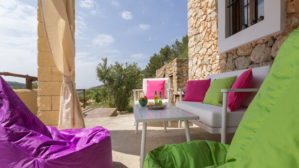 An outdoor seating area with a stone wall, a white sofa with colorful cushions, and a purple bean bag. - Typical Ibizan country house with private pool in a beautiful protected environment! (ISLAS BALEARES)