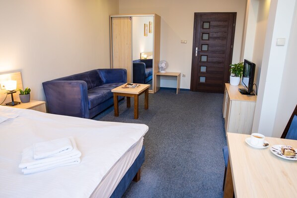 Habitación de hotel con cama, sofá azul, pequeña mesa con lámpara, escritorio con computadora y puerta con espejo.
