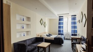 A compact hotel room with a sofa, a single bed, a small table, and a desk with a chair. There are shelves with decorative items and a window with curtains. - Hotel Kardamon (Tarnow)