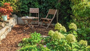 A garden with a wooden table and two chairs, surrounded by various plants and a stone wall. - Experience with us the Harz im Glück! Vacation for two in Wernigerode! (Wernigerode)