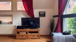 A living room with a television on a wooden stand, a grey sofa, a red curtain, and a window with a view of buildings and trees. - Experience with us the Harz im Glück! Vacation for two in Wernigerode! (Wernigerode)