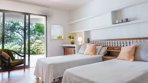 Quarto de hotel com duas camas, cabeceira de madeira, uma mesinha com abajur e vista para o verde através de uma porta de correr.