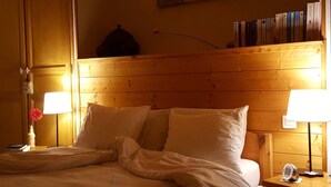 Une chambre d’hôtel avec une tête de lit en bois, une table de chevet avec une lampe, un petit vase avec une fleur et une bibliothèque remplie de livres.