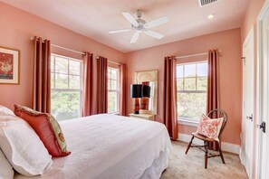 Une chambre à coucher comprenant un lit, une chaise, une table de chevet, un ventilateur de plafond et une fenêtre avec des rideaux.