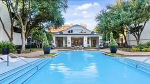 Una grande piscina rettangolare con superficie d'acqua cristallina e azzurra, circondata da un solarium piastrellato. Ai lati della piscina ci sono due grandi vasi con piante, e sullo sfondo un edificio con tetto grigio e pareti bianche.
