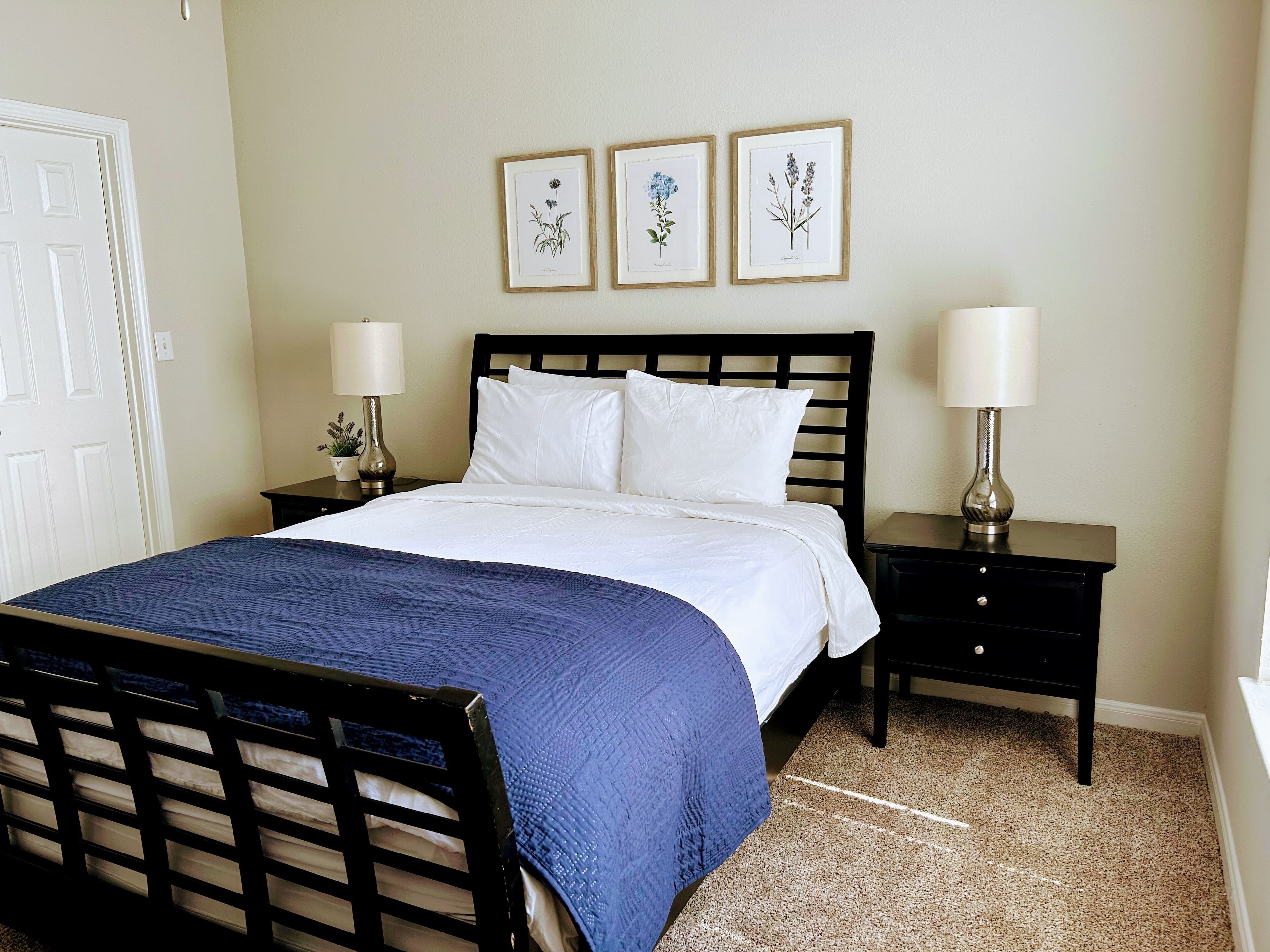 A neatly made bed with white linens and a blue blanket, flanked by two bedside tables with lamps, in a room with a beige carpet and a door visible to the left.