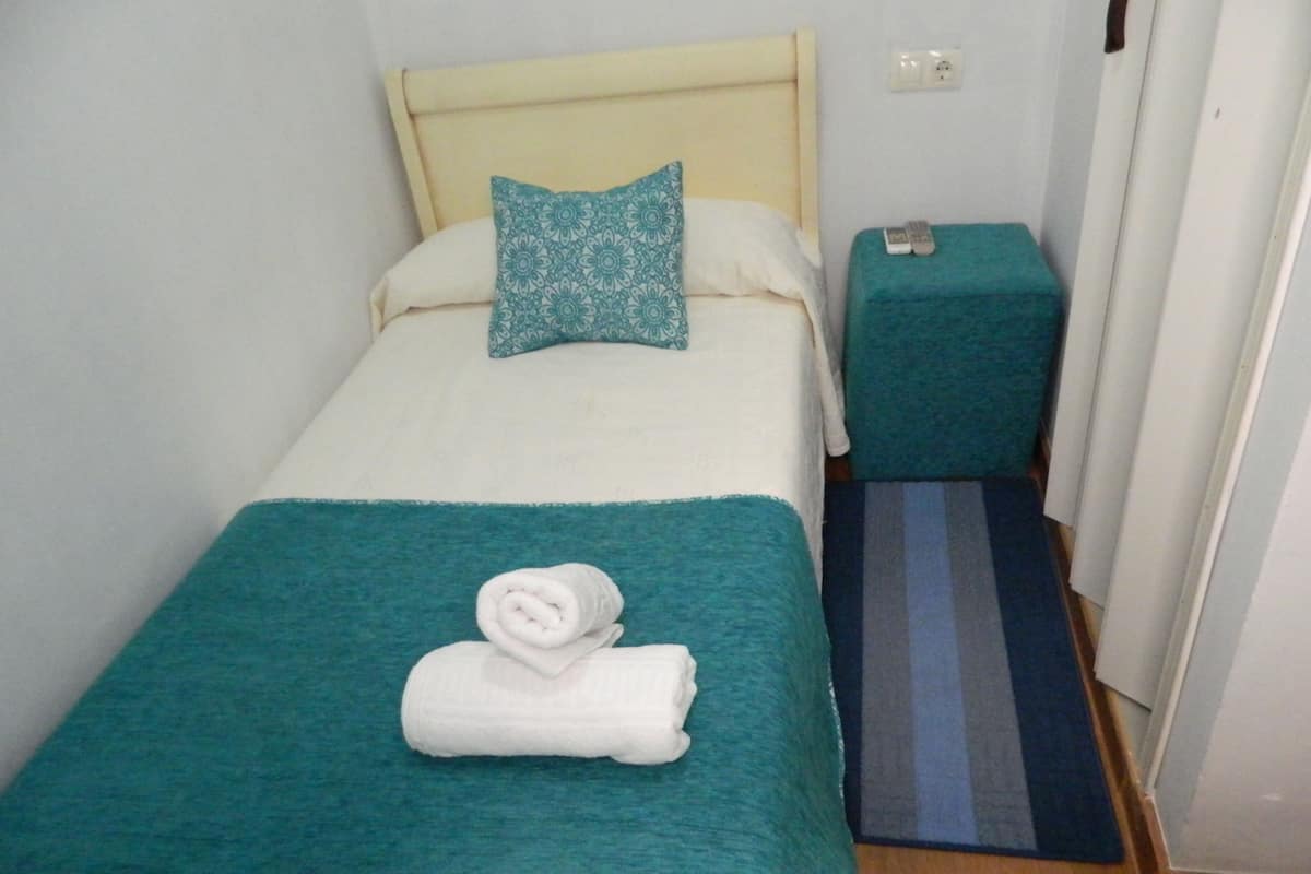 Un llit individual amb un edredó verd mar i una tovallola blanca arrotllada a sobre. A sobre d'una tauleta de nit a joc, de color verd mar, hi ha un comandament a distància.