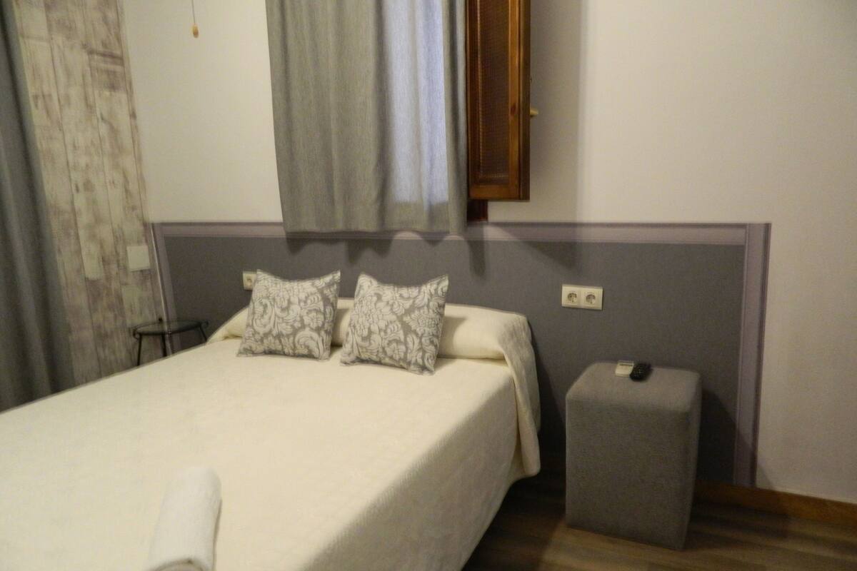 Un llit ben fet amb dos coixins, una taula de nit amb un comandament a distància i un petit otoman.