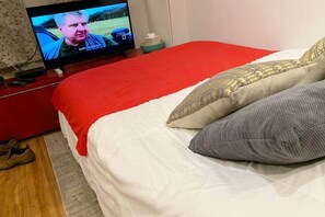 Un dormitorio con televisión sobre un mueble, una colcha roja y una cama blanca.