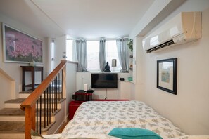 A bedroom with a bed, a TV, a window with curtains, a staircase, and an air conditioning unit.