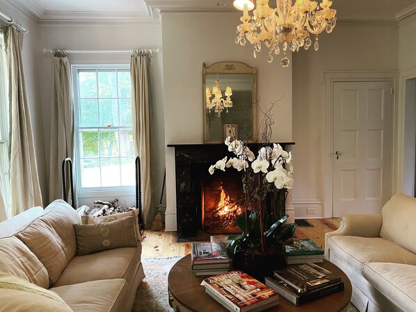 A living room with a fireplace, two sofas, a coffee table with books, and a chandelier. - Magnificent historic mansion on 12 acres with flower gardens. Pet friendly (Hopewell Junction)