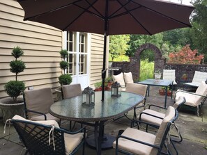 Eine Terrasse im Freien mit einem runden Glastisch, beigefarbenen Sitzgelegenheiten, einem großen Sonnenschirm und einem Backsteinbogen.
