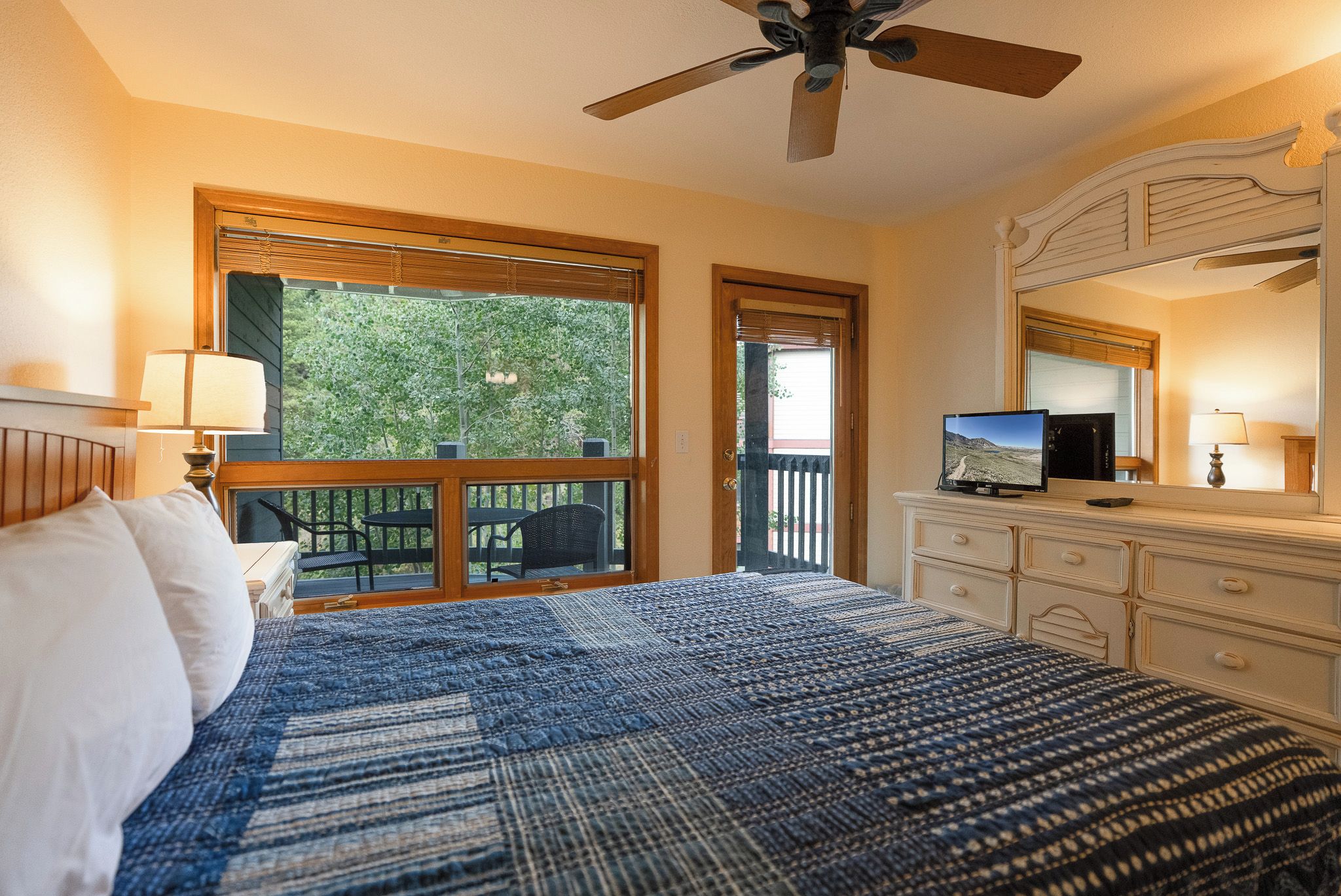 Une chambre à coucher avec un lit, un téléviseur posé sur une commode, un ventilateur de plafond et une fenêtre donnant sur un paysage verdoyant.