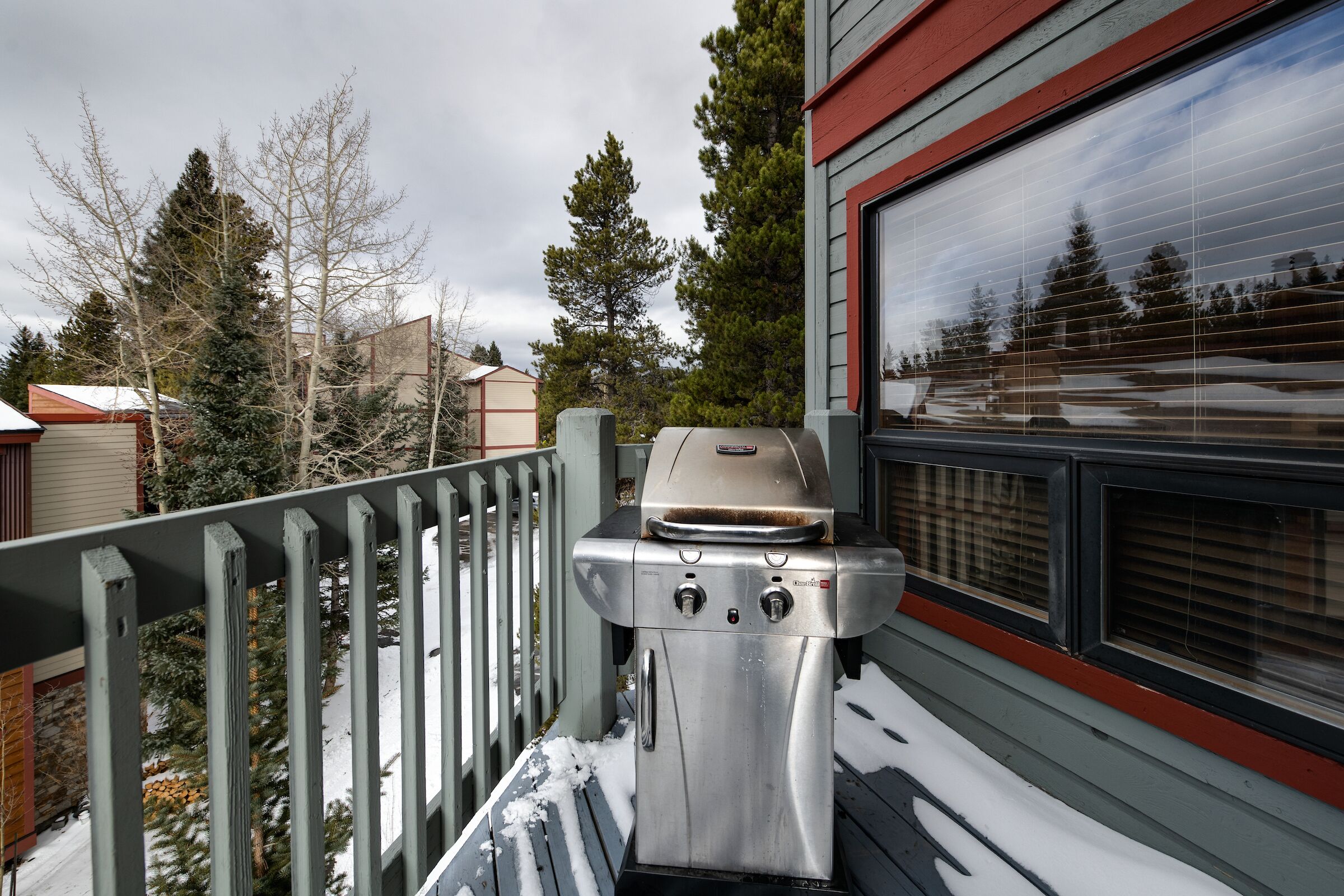 A stainless steel grill on a balcony with snow on the ground, surrounded by trees and a building with a window.