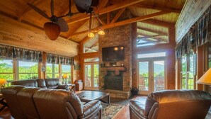 A log cabin interior with a fireplace, leather sofas, and large windows. - 4BR cabin with hot tub, game room, fire pit, and mountain views. (Sautee Nacoochee)