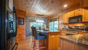 A kitchen with wooden cabinets, a stone wall, a bar area, and a large window. - 4BR cabin with hot tub, game room, fire pit, and mountain views. (Sautee Nacoochee)