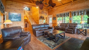 A log cabin interior with a fireplace, leather sofas, a dining table, and a staircase leading to a loft. - 4BR cabin with hot tub, game room, fire pit, and mountain views. (Sautee Nacoochee)