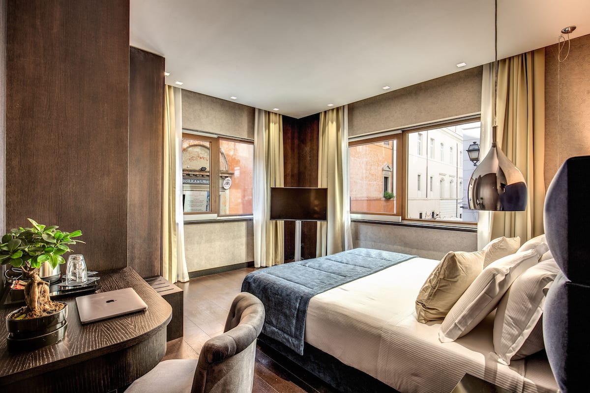 A modern hotel room with a bed, a desk with a laptop, a chair, and a potted plant.