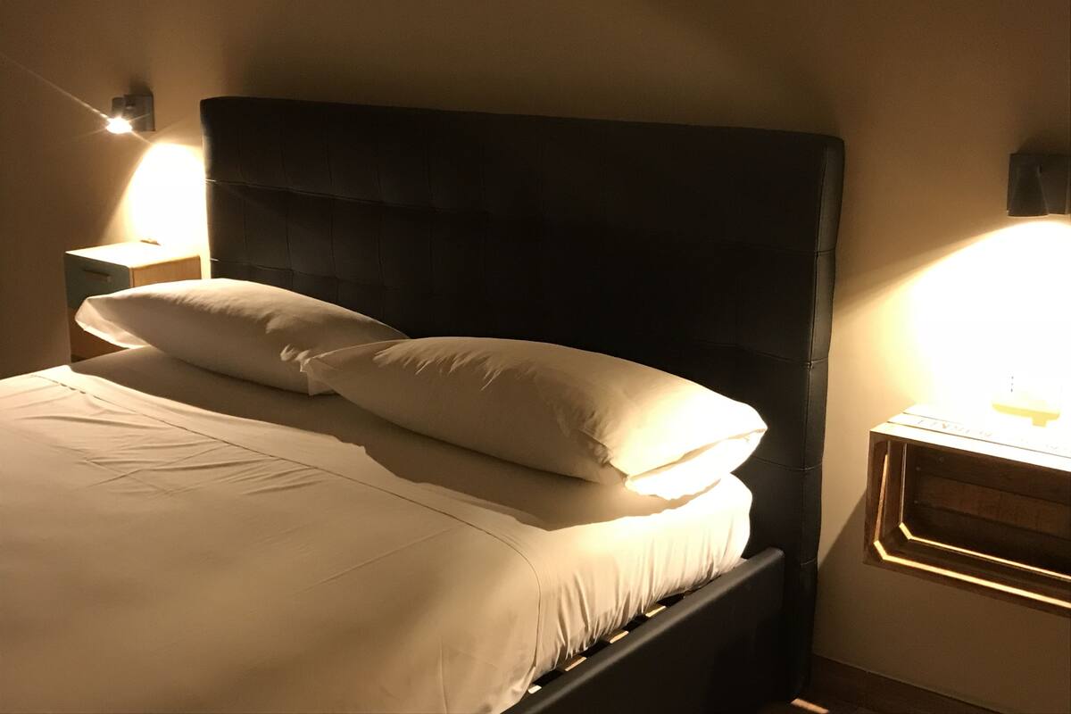 Un lit avec une tête de lit sombre, deux oreillers et une lampe de chevet posée sur une table de nuit en bois.