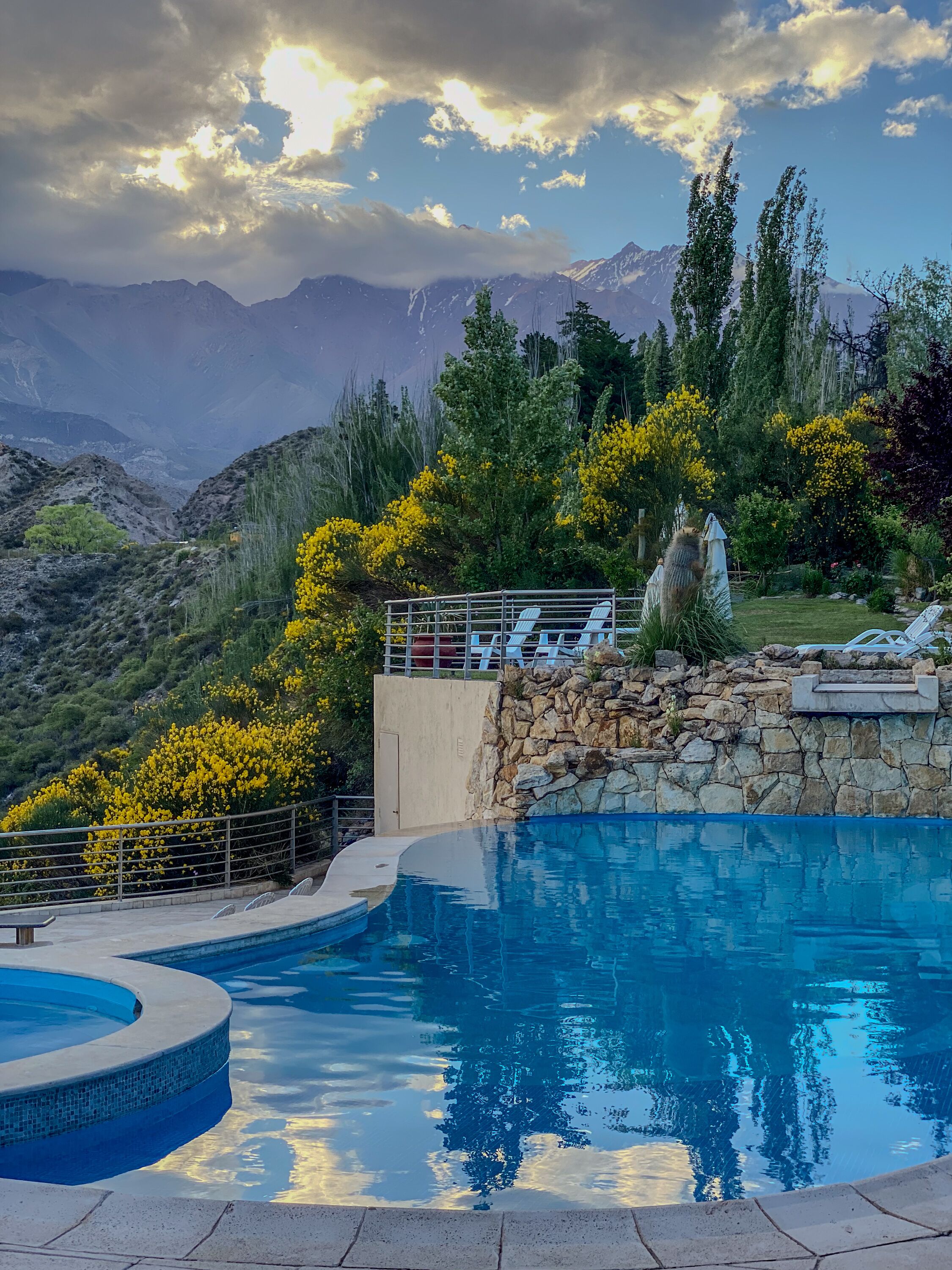 Una piscina con pared de piedra y sillones de descanso, rodeada de vegetación y montañas al fondo.