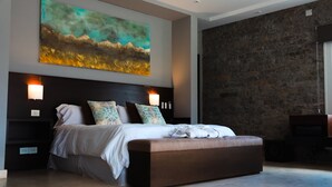 A modern bedroom with a large bed, a headboard with wall sconces, a stone accent wall, and a painting above the bed. - El Carmelo Mountain Lodge (Potrerillos)
