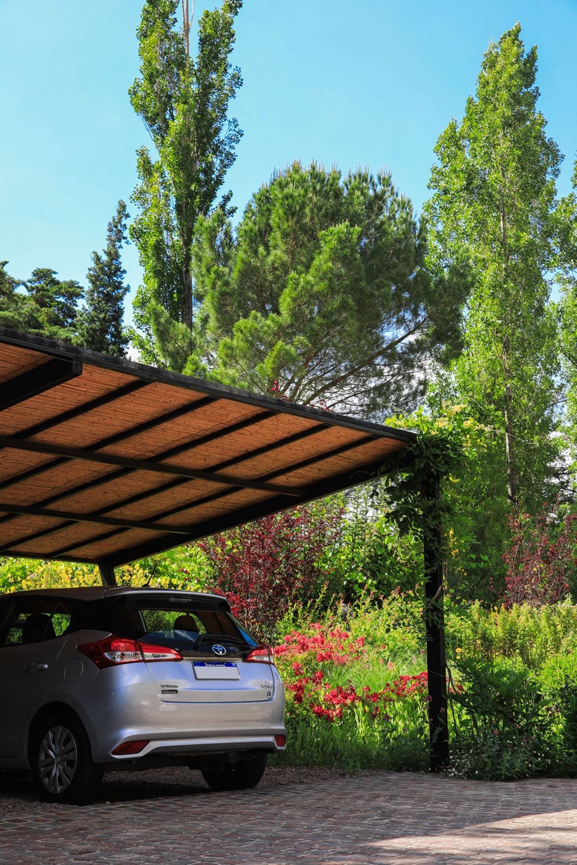 A silver SUV parked under a wooden pergola in a garden with trees and flowering plants.